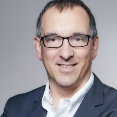 Markus Georg trägt kurzes, dunkles Haar, eine schwarze Brille und einen dunklen Anzug mit weißem Hemd.