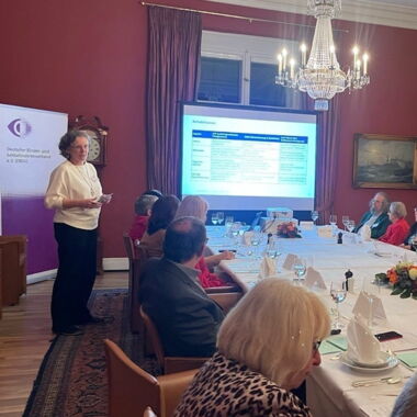 Beatrix Seeliger steht im hinteren Teil des Raumes vor der Projektionswand und dem DBSV Aufsteller und führt das Gespräch mit Christel Pildner und Roswitha Röding